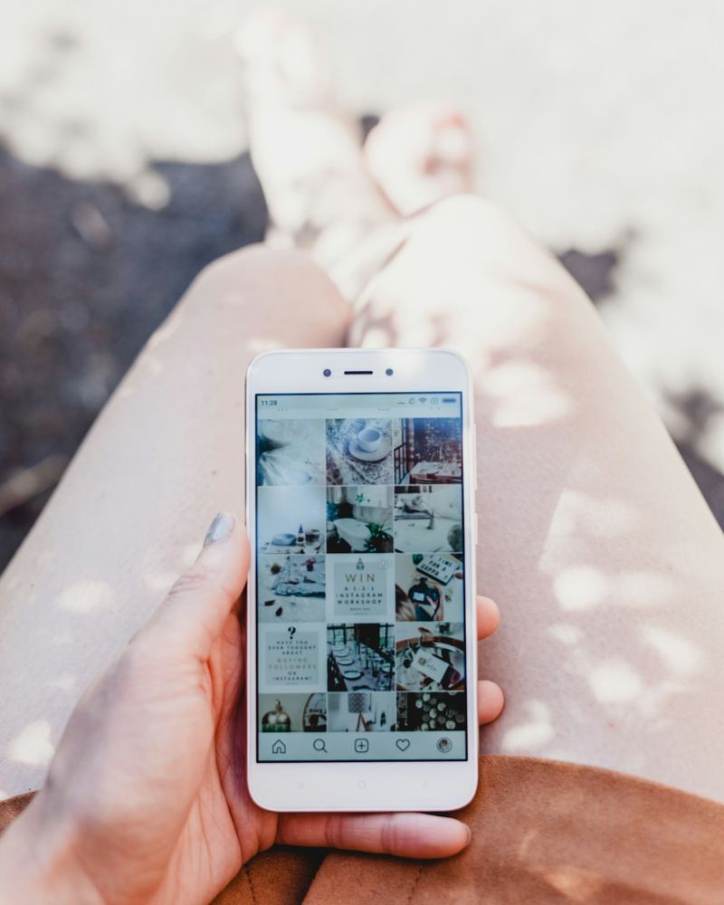 A person holding a white smartphone showing an Instagram feed in bright sunlight