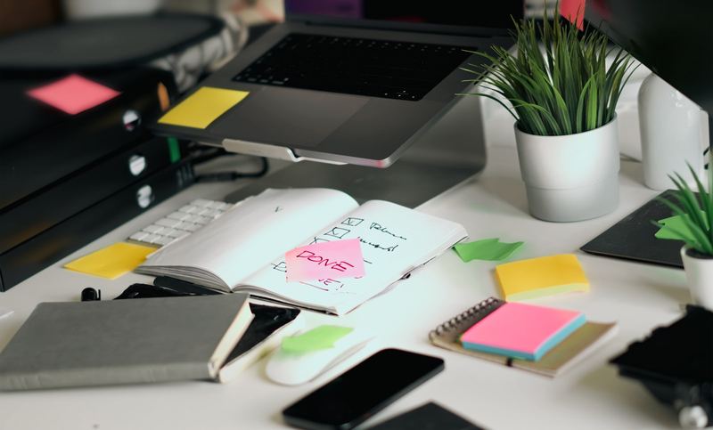 A cluttered desk with a laptop, a smartphone, an open notebook and scattered sticky notes
